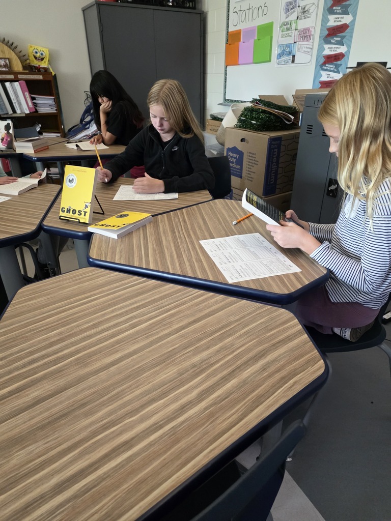 CMS students participate in a book tasting activity, reading sample books and completing worksheets at classroom desks. Two students in the foreground examine books, including Ghost, while another student reads in the background.