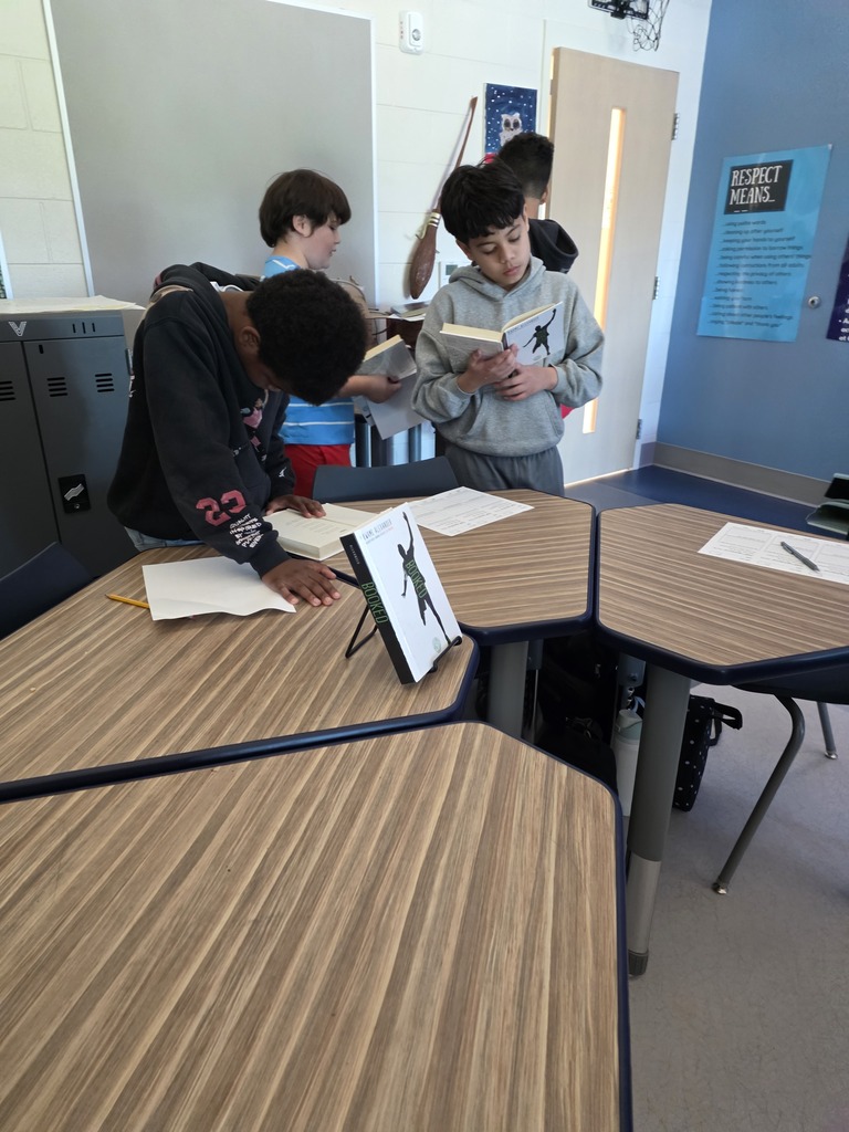Four CMS students explore books during a classroom book tasting activity. One student leans over a desk to examine a book while another reads Booked. Other students browse books nearby.