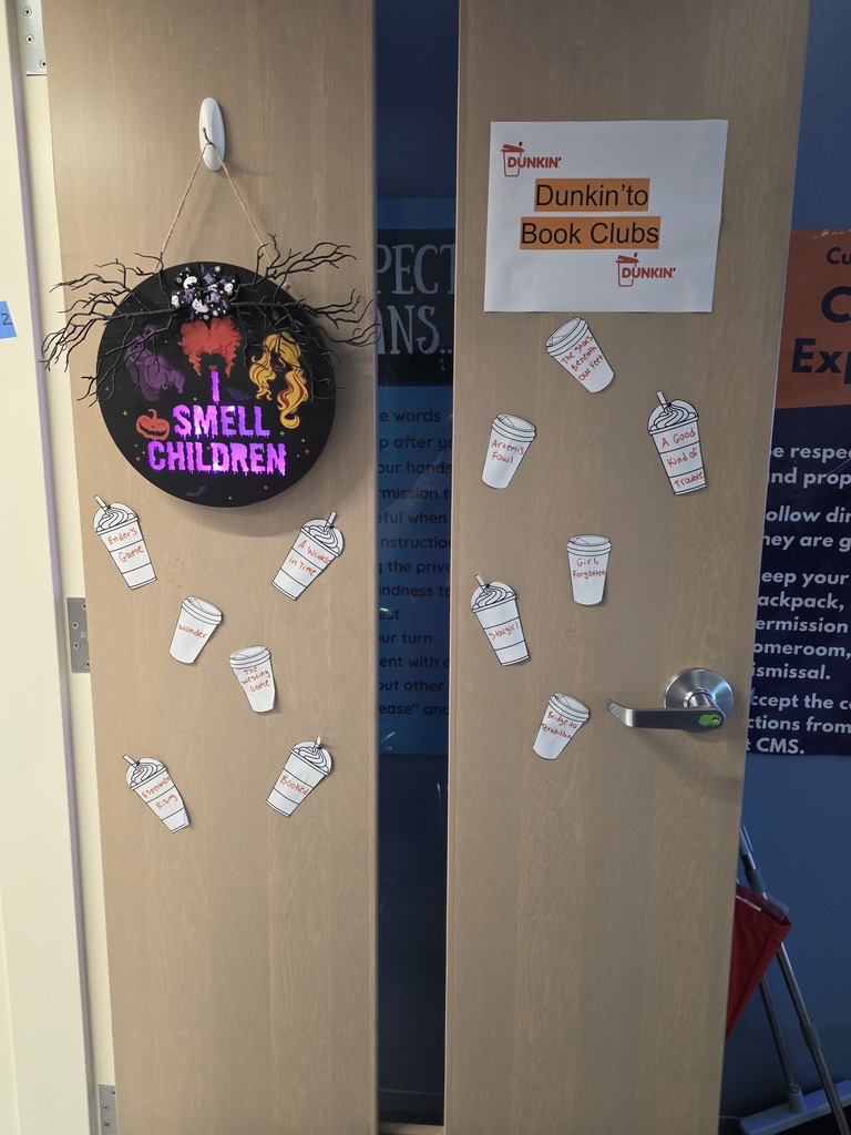 A classroom door decorated for a book tasting activity features a sign reading “Dunkin' to Book Clubs.” Paper coffee cups displaying different book titles are attached to the door, along with a Halloween-themed “I Smell Children” decoration.