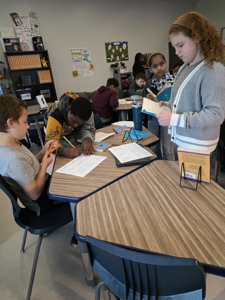 CMS students explore different books during a classroom book tasting activity. Some students read while others complete worksheets to help select their book clubs for the quarter. Books are displayed on stands throughout the classroom.