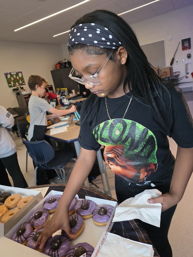 A CMS student selects a decorated donut from a box during a celebration following the book tasting activity. The donuts feature purple frosting, chocolate drizzle, and cookie decorations resembling spiders, with additional glazed donuts nearby.
