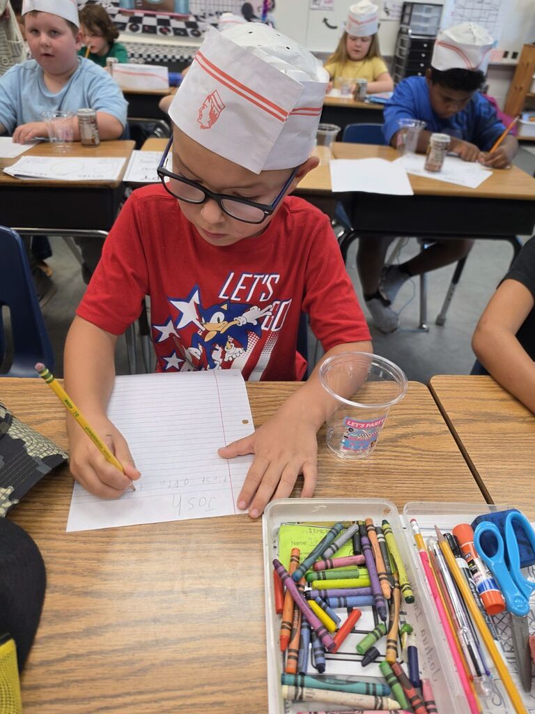 Student seated at a desk, wearing chef hat and glasses, writing on paper with pencil.