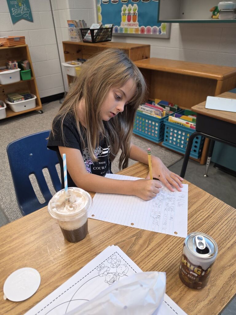 Girl at table in classroom, writing on paper, pencil in hand, a cup of drink, a soda can.