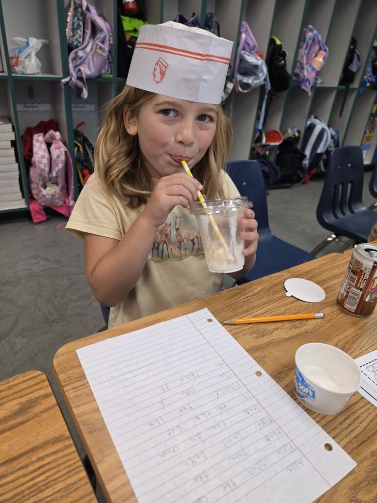 Child in chef hat drinks from cup through straw, seated at a table, paper, pencils, and cup.