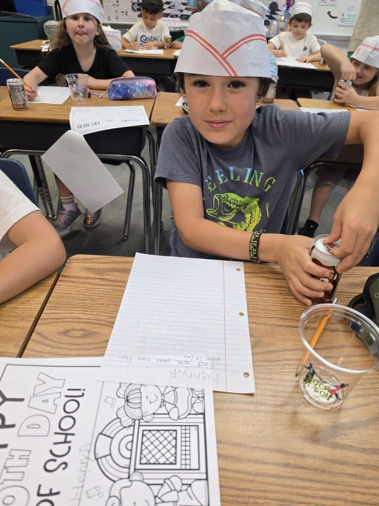 Students sit at desks, wearing paper hats, with one holding a bottle and a cup. Papers with drawings and text are on the desk.