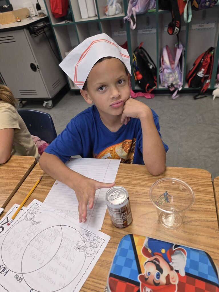 Boy in chef hat sits at table with papers, pencil, can, and glass. Empty chairs, backpacks, and lockers behind him.
