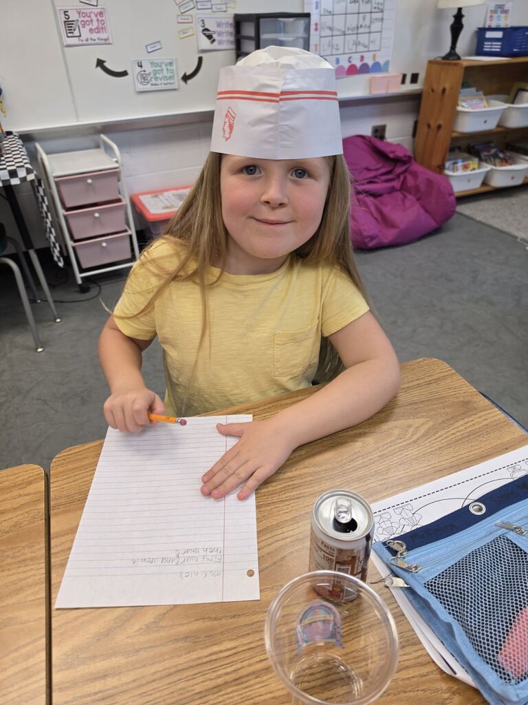 Child in chef hat writing at desk, paper, pencil, soda can, plastic cup, open pencil case, rug, shelves in background.