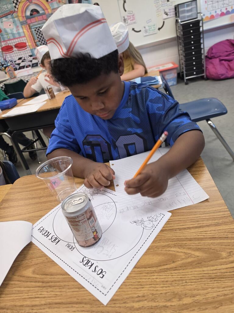 Child wearing chef's hat, writing on paper with a pencil at a desk, a can and cup nearby.