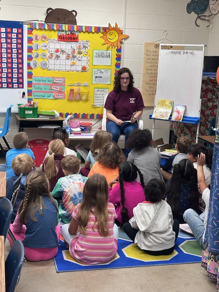 Woman reading to students in circle, seated on chair, carpeted floor, colorful classroom bulletin board.