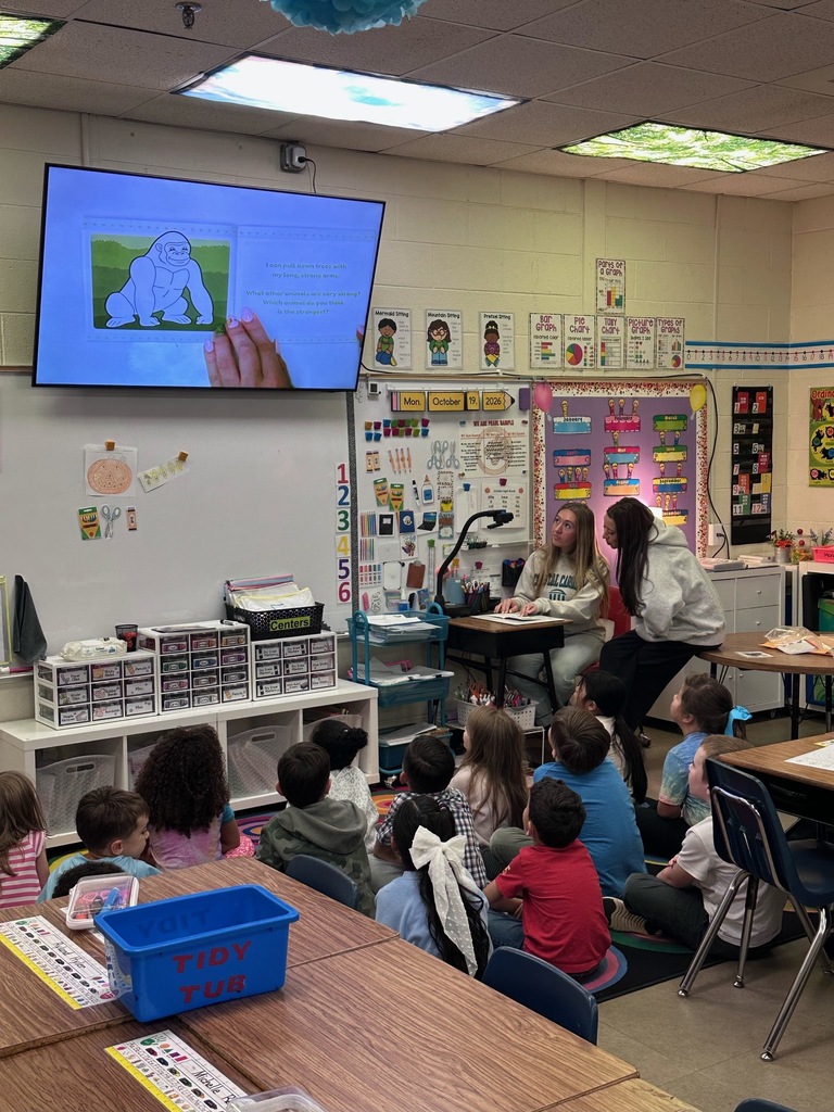 Students sit in a semicircle in front of a teacher on a raised platform. A monitor displays a cartoon image.