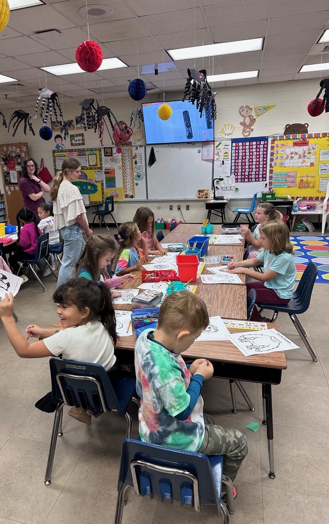 Kids sitting at tables in a classroom, some holding paper and colored pencils.