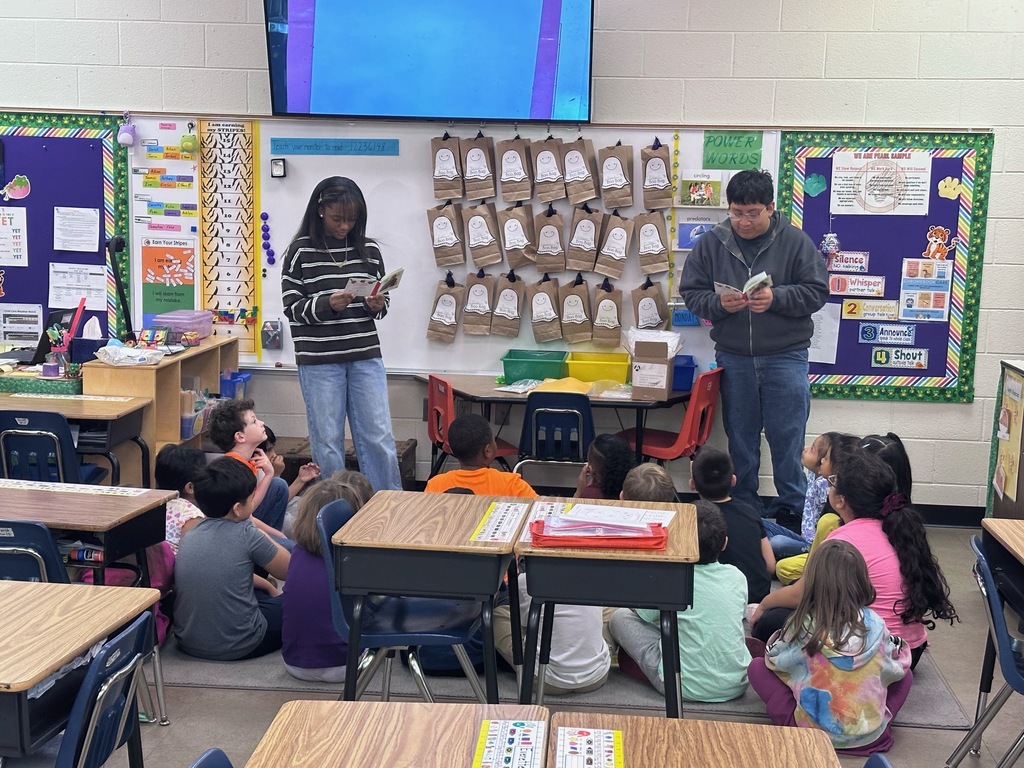 Adult and child read aloud to students sitting in a classroom, tables and chairs in background.