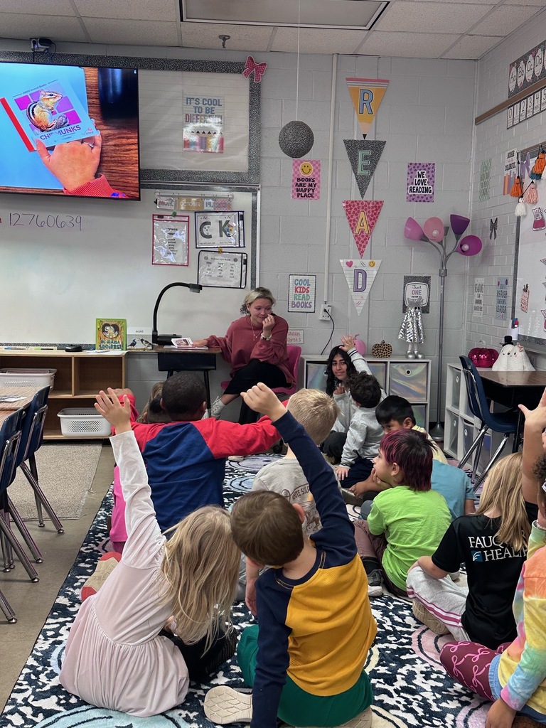 Students sit cross-legged on a rug in a classroom; a teacher sits on a chair.