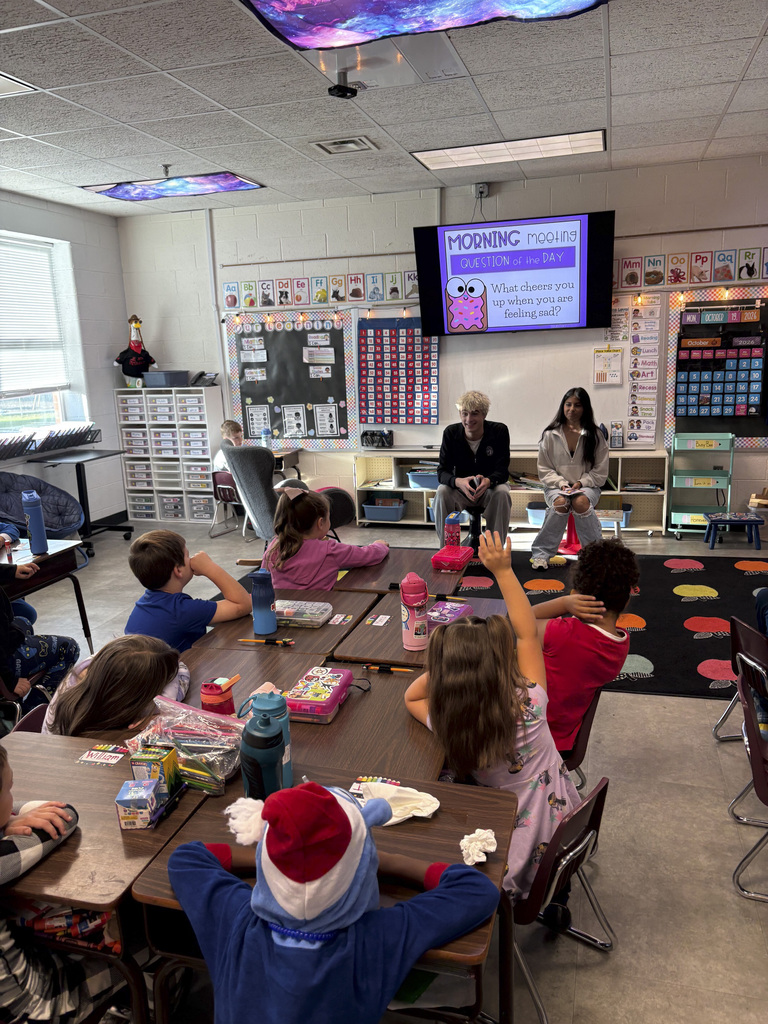 Classroom with students seated at tables, two adults at the front, screen projector, and colorful rug.