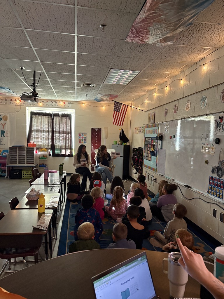 A classroom with students sitting around a table, facing a whiteboard, a flag, and a ceiling fan.