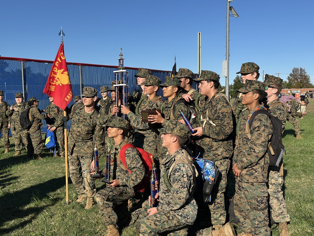 Military cadets holding trophies and flags on a grass field with a fence in the background.