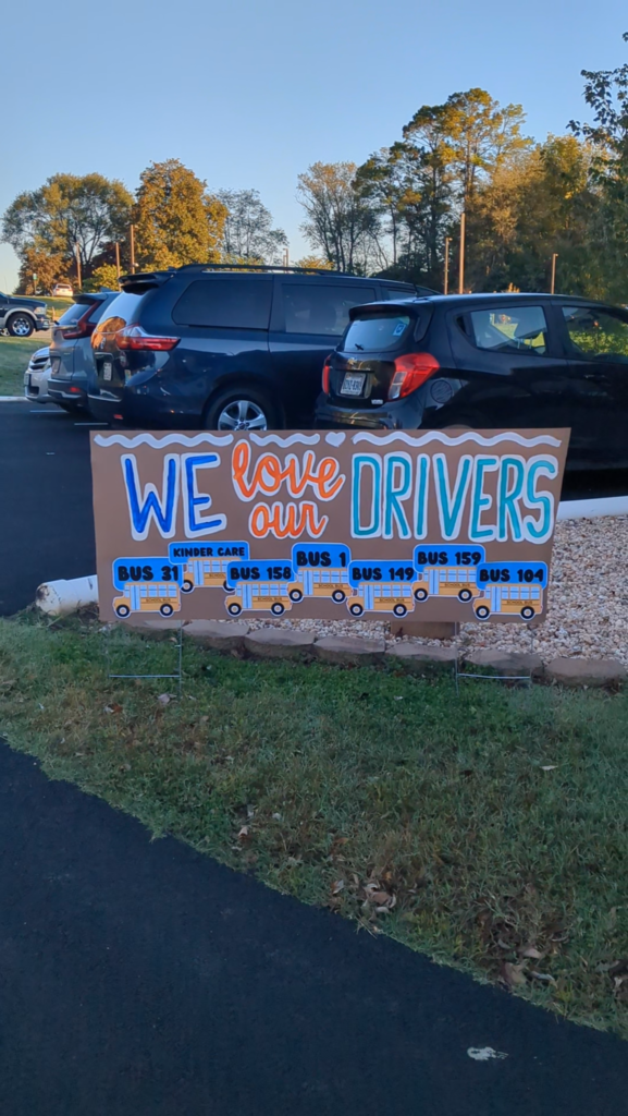 A sign reading "WE love our DRIVERS" stands on grass. Behind it, cars park in a lot. Trees line the edge of the lot.