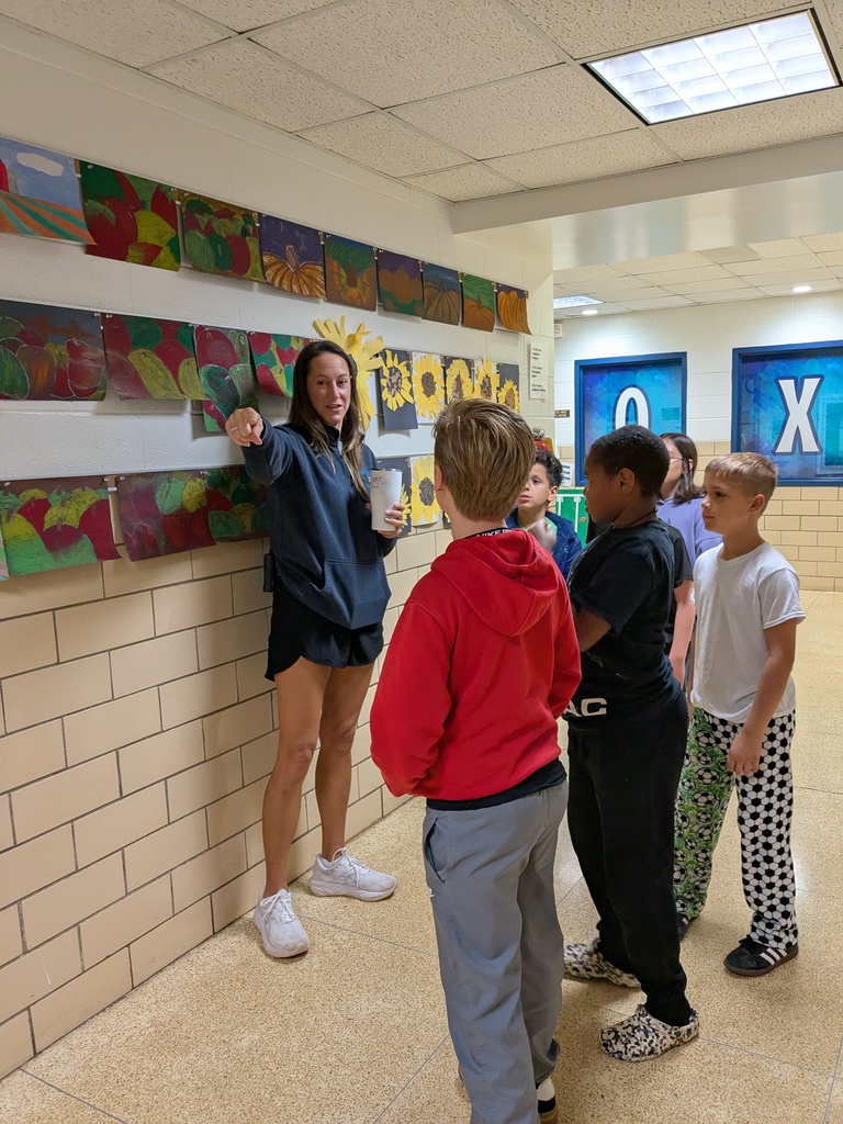 Woman with cup standing near wall with colorful artwork, talking to group of children.