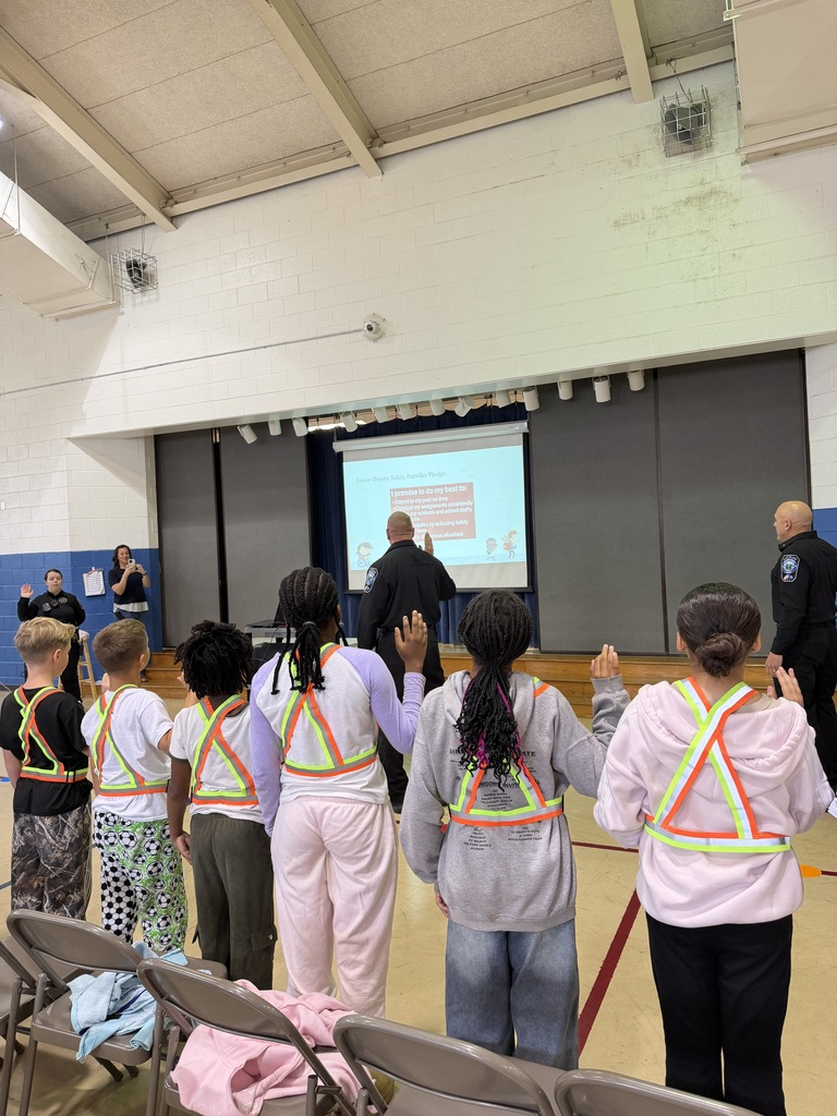 Kids in reflective vests stand in a row in a gym as an adult gives a presentation on a screen.