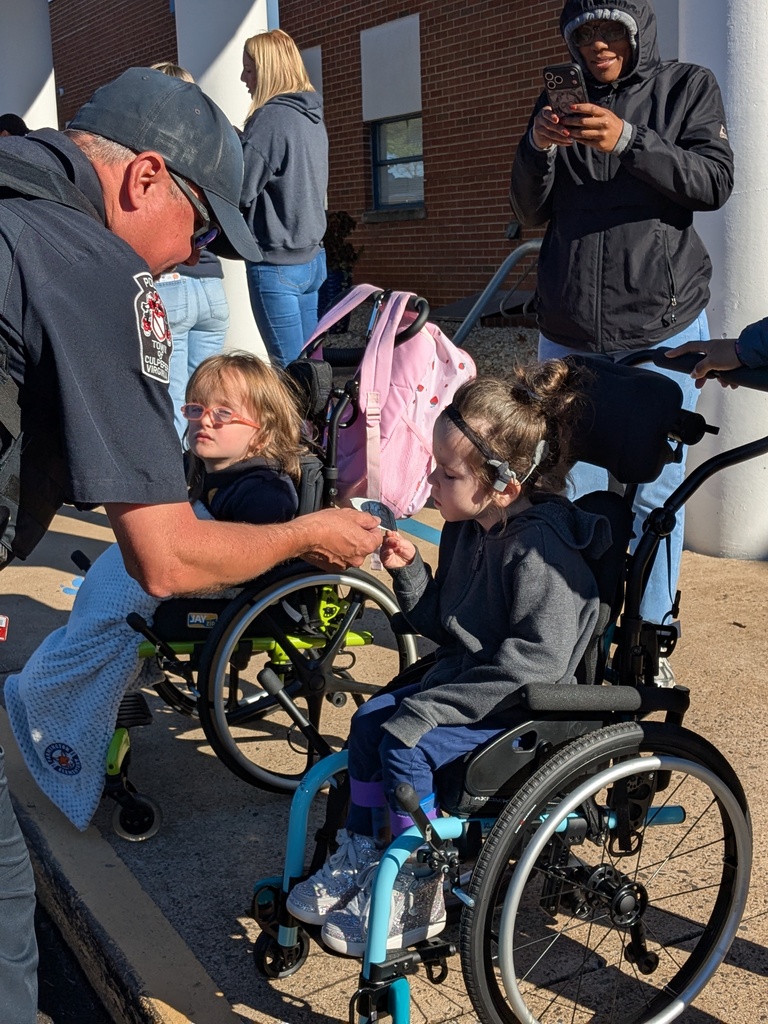 Man helps child in wheelchair with glasses. One child in pink backpack stands nearby. Two people in background.