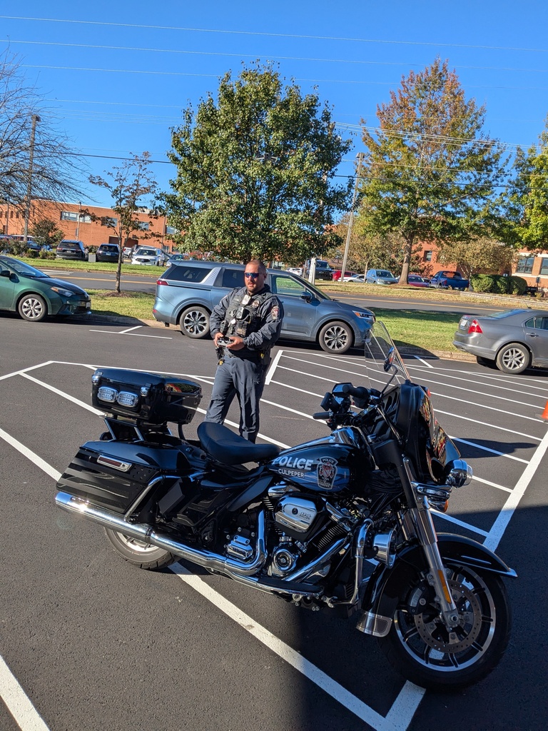 A person stands behind a black motorcycle in a parking lot with parked cars, trees, and buildings.