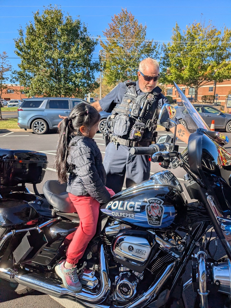 A child sits on a motorcycle while a police officer stands nearby. Trees and parked cars are in the background.