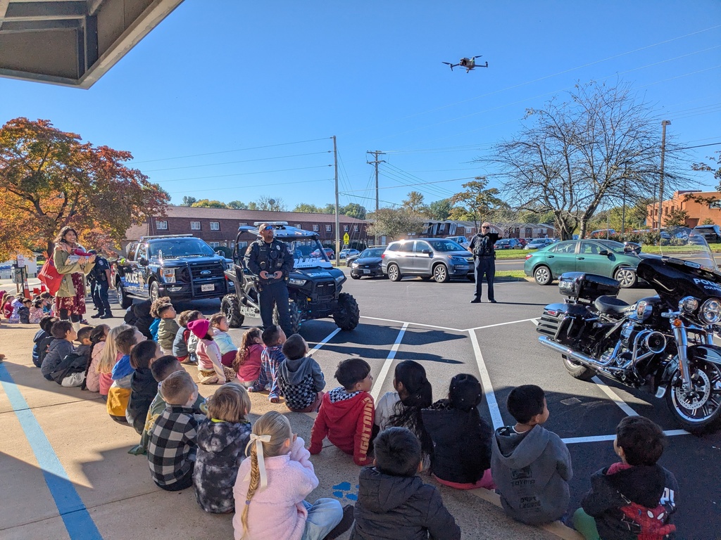 Children sitting on a sidewalk watch a drone fly over a parking lot with cars and police vehicles.