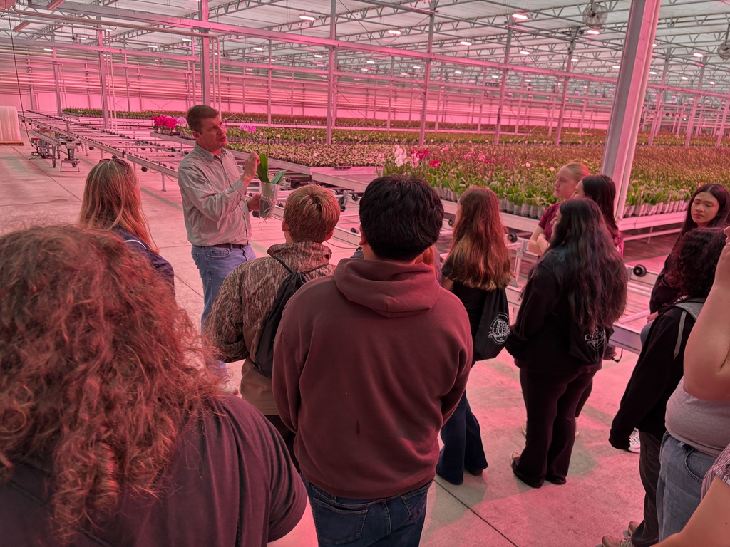 Several students in a greenhouse observe plants as a man stands and speaks to the group.