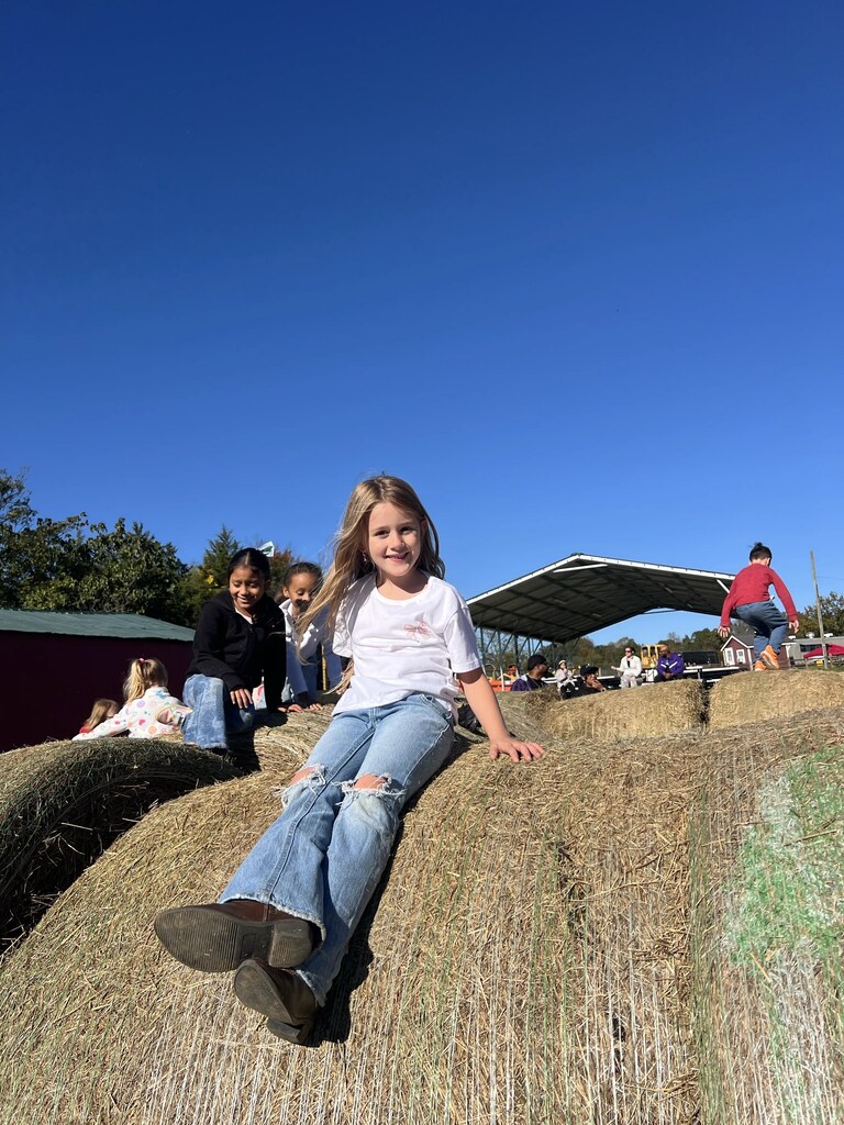 Four kids sit atop a large hay bale under a blue sky; one child leans forward with hands on knees.
