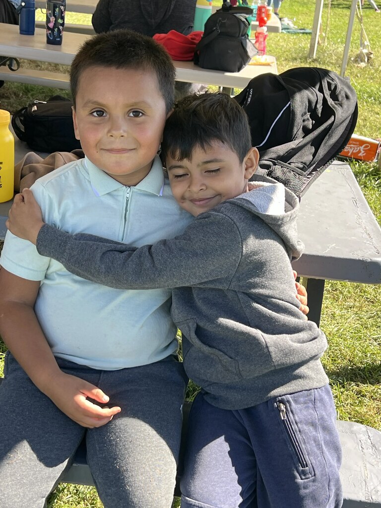 Two boys in casual clothes sitting on a bench outside, one hugging the other from behind.