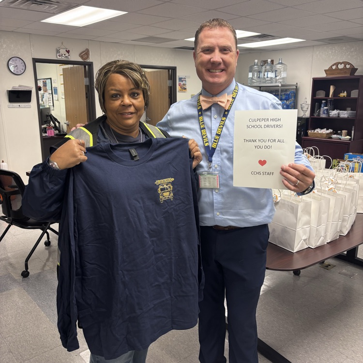 A man and woman hold a blue T-shirt and paper in an indoor room. A desk with bags is behind them.