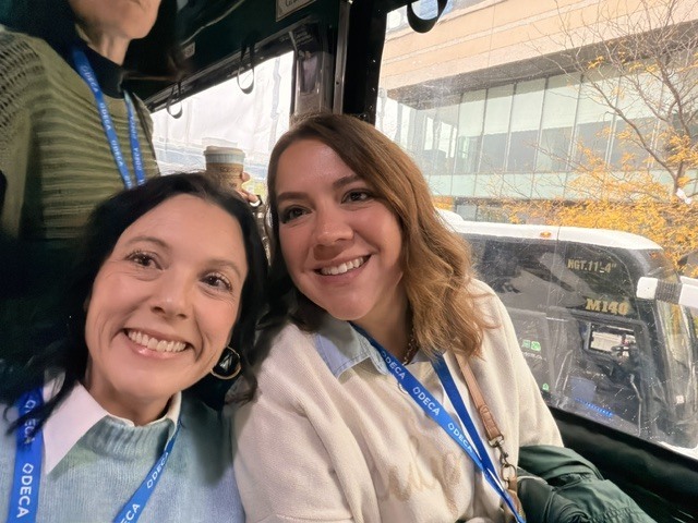Two women in green lanyards sit on a bus, smiling and facing the camera, with a glass window behind them.