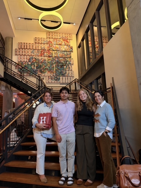 Four adults stand on stairs in a lobby. Two women on the sides hold bags. One man wears a pink shirt.