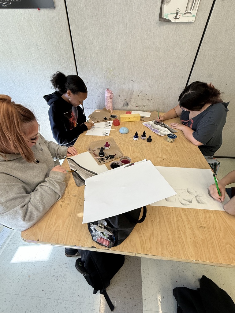 Four students work at a table, seated in chairs. Three hold pens and paper. One has a bag on the floor.