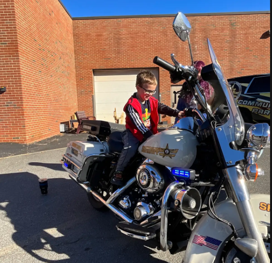 A child sits on a police motorcycle with a flag on the side, in front of a brick building.