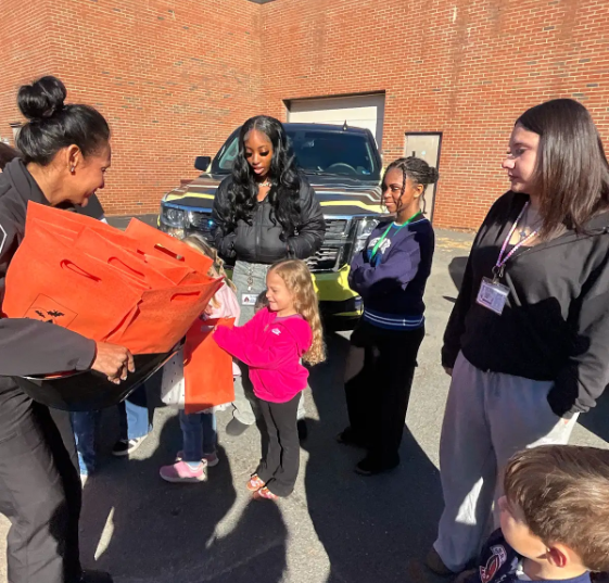A woman hands orange bags to a young girl. A few adults and children stand nearby. A brick building is in the background.