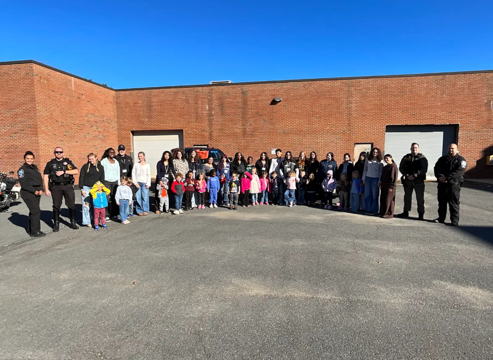 A large group of people, including adults and children, stands in front of a brick building on a sunny day.