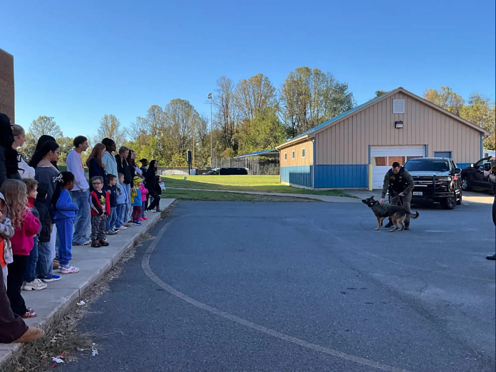 Group of people in winter clothing standing on sidewalk, watching a police officer and dog.