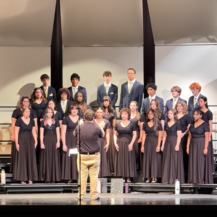 A group of students in formal attire stand on a stage in rows, singing. A man stands before them.