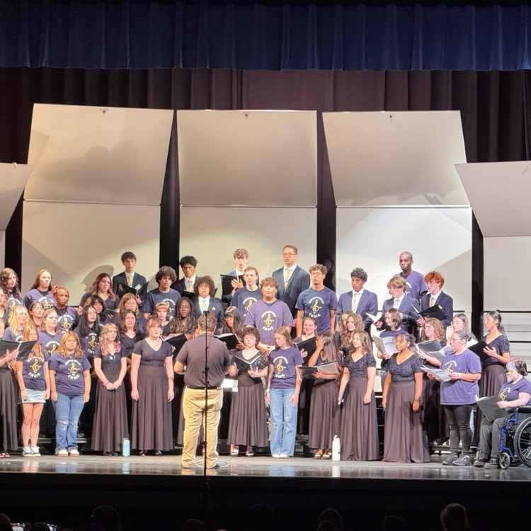 Group of students wearing purple shirts stand on stage with music stands in front of them.