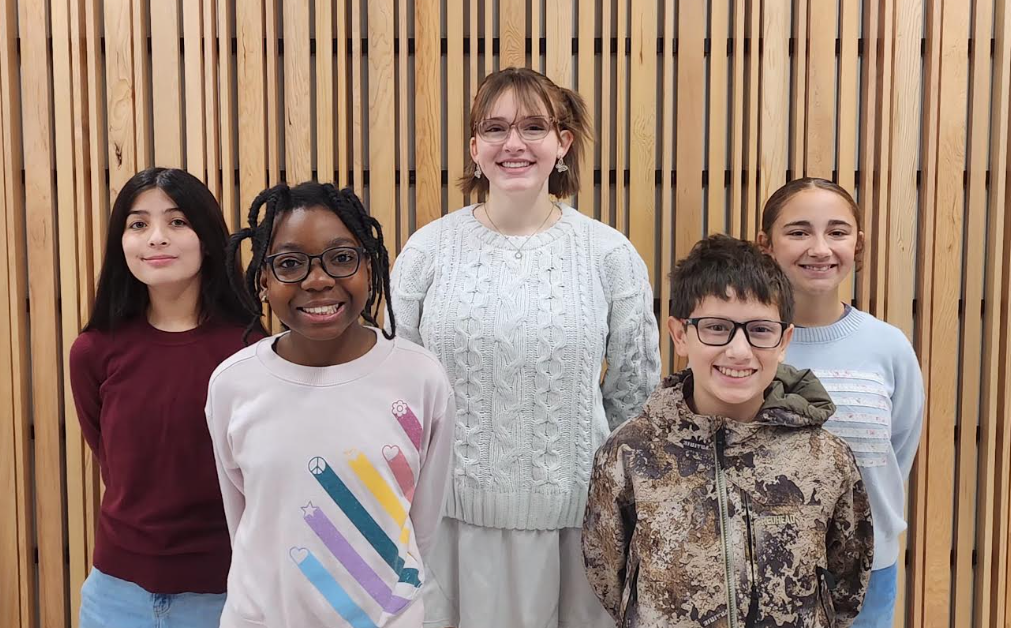 Five CMS Student Council Association (SCA) leaders pose together in front of a wooden slat wall. Pictured from left to right are Bianca Do, 8th Grade Representative, wearing a burgundy sweater; Divine Nteham, 6th Grade Representative, wearing a light pink sweatshirt; Sutton Brink, President, wearing a white cable-knit sweater; Frank Alonso, 7th Grade Representative, wearing a camouflage hoodie; and Adriana Saylor, Vice President, wearing a light blue sweatshirt. All five students are smiling at the camera.