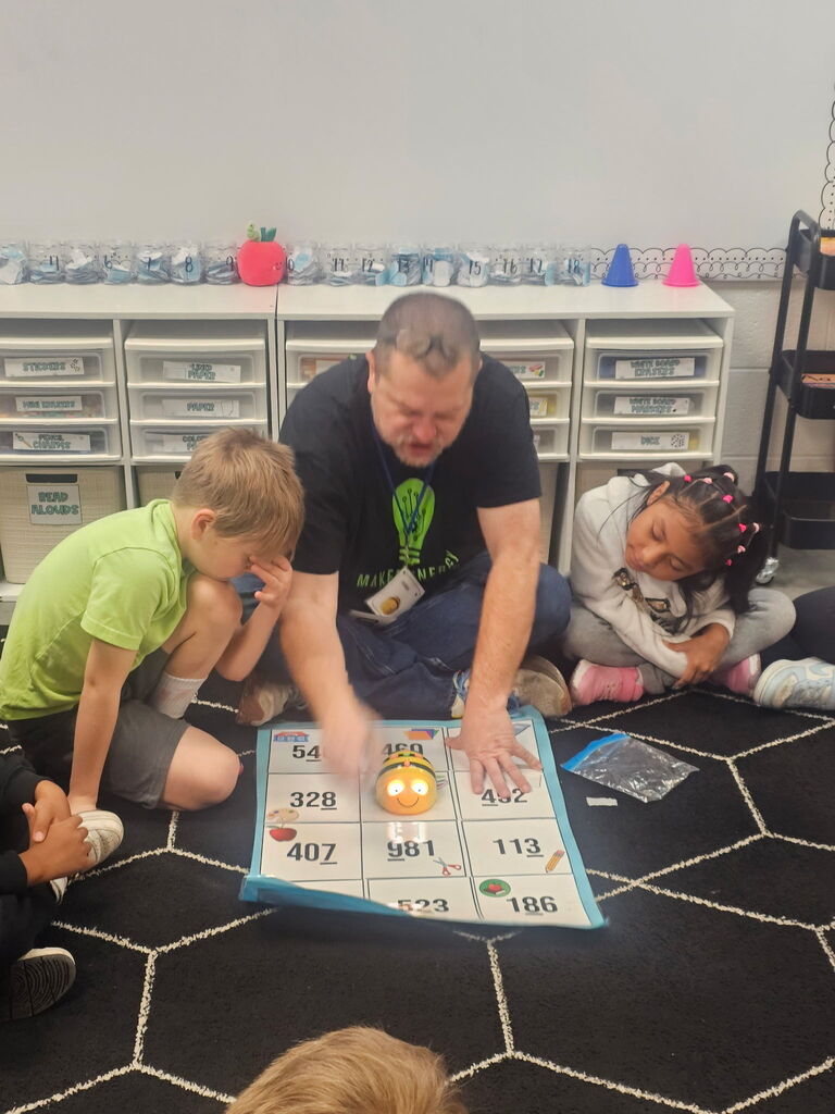 Teacher helps students with a numbered game board, a small robot, and small plastic containers.