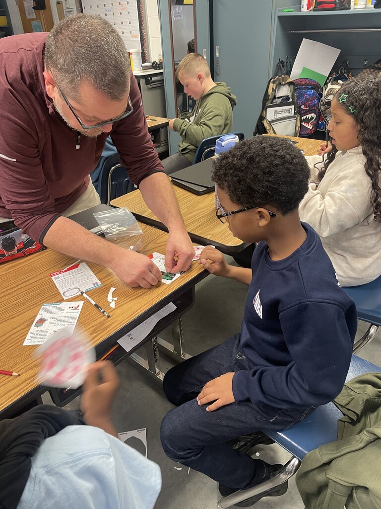 Adult helps a student with a card. Other students and an adult sit in the background.