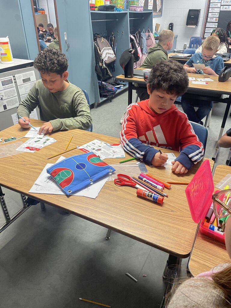 Two young boys at tables with coloring materials and paper; other students seated in background.