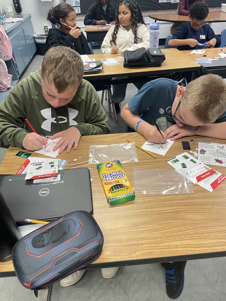 Students seated around a table, two wearing glasses, writing on paper with pens and markers.