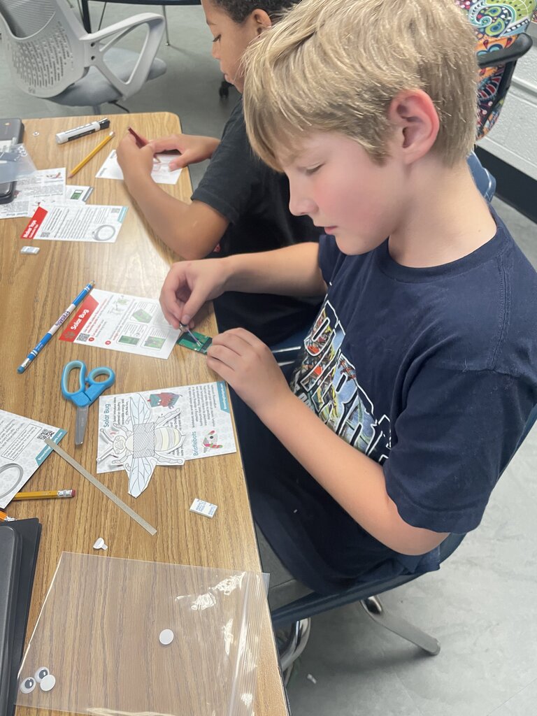 Two children sit at a table. One child uses scissors to cut paper. The other child has a circuit board.