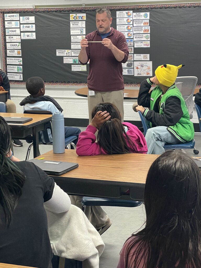 A teacher stands at a desk with a ruler, showing it to students seated at tables. One student rests head on the desk, another in a green vest looks at the teacher.