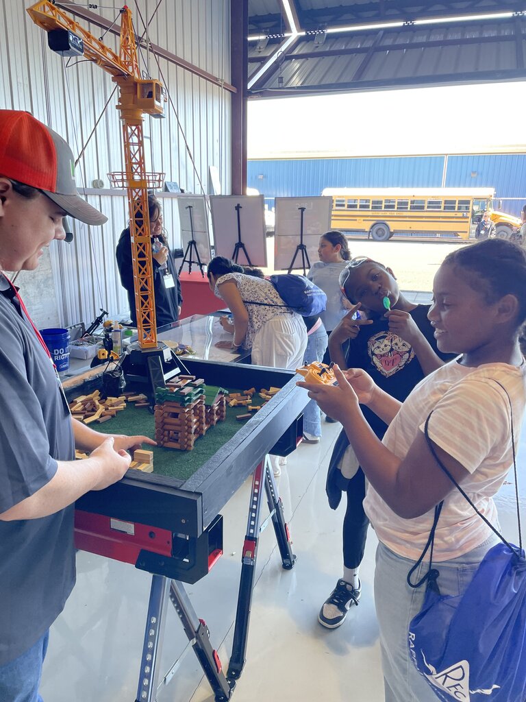 A boy eats a small sandwich while a man adjusts a large model building. Nearby, a girl holds a camera.