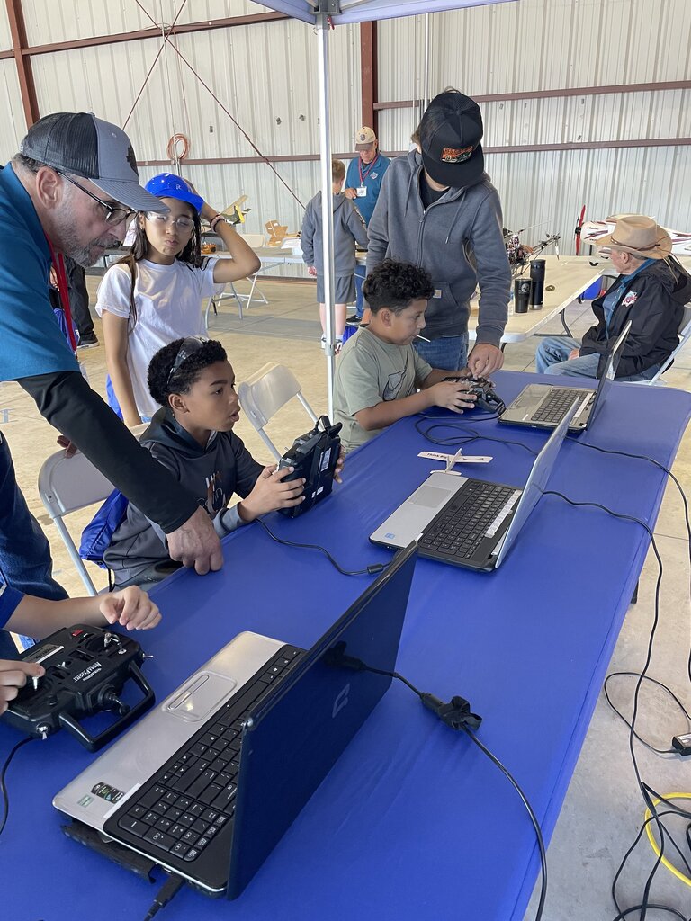 Several children, seated at a table under a tent, operate electronic devices with a man nearby.