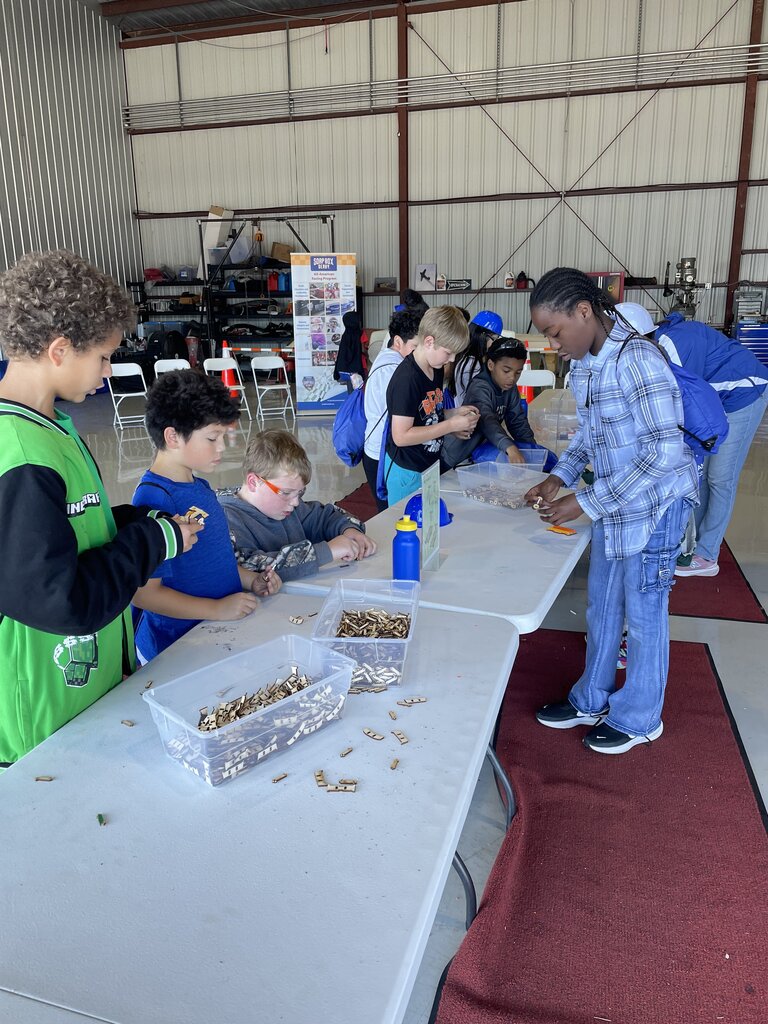 Several children work at tables sorting small objects in clear containers inside a hangar.